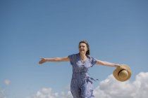 Contenu femme adulte avec cheveux soufflants et en robe de soleil marchant avec chapeau de paille à la main sur fond de ciel nuageux — Photo de stock