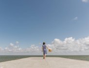 Back view of adult woman in straw hat and sundress on empty concrete quay on cloudy day — Stock Photo