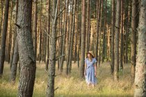 Happy adult woman in straw hat and sundress standing in forest among coniferous trees in golden sun ray — Stock Photo