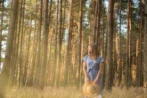 Счастливая взрослая женщина в соломенной шляпе и сарафане, стоящая в лесу среди хвойных деревьев в золотом луче солнца — стоковое фото