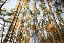 Mulher adulta feliz em chapéu de palha e sundress em pé na floresta entre árvores coníferas em raio de sol dourado — Fotografia de Stock