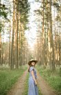 Back view of adult woman in straw hat and sundress walking along forest road between pines at sunny day — Stock Photo
