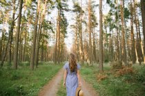 Back view of carefree adult woman in straw hat and sundress walking along forest road between pines at sunny day — Stock Photo