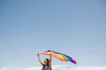 Vista lateral de mujer confiada adulta en vestido casual llevando bandera de color arco iris por encima de la cabeza en el día ventoso - foto de stock