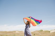 Вид на взрослую уверенную женщину в повседневной одежде с радужным флагом над головой в ветреный день — стоковое фото