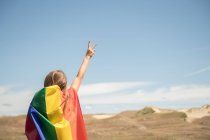 Vue arrière de la femme confiante adulte en robe décontractée portant drapeau de couleur arc-en-ciel au-dessus de la tête le jour venteux — Photo de stock