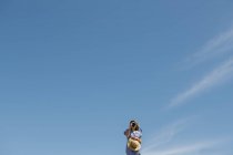 Donna in cappello di paglia e vestito con la macchina fotografica, in piedi in soleggiata giornata ventosa con cielo azzurro chiaro su sfondo — Foto stock