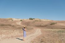 Adult woman in straw hat and dress with camera taking photo on unpaved road among dry dusty field on sunny day — Stock Photo