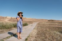 Adult woman in straw hat and dress with camera standing on country path — Stock Photo