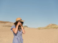 Femme adulte en chapeau de paille et robe avec caméra prenant des photos sur la dune de sable de la plage dans une journée ensoleillée — Photo de stock