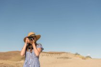 Femme adulte en chapeau de paille et robe avec caméra prenant des photos sur la dune de sable de la plage dans une journée ensoleillée — Photo de stock