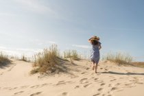 Mujer adulta en vestido casual y sombrero de paja caminando pasar un buen rato en la duna de arena en el día de verano caliente - foto de stock