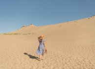 Donna adulta in abito casual e cappello di paglia che cammina divertendosi sulle dune di sabbia nella calda giornata estiva — Foto stock
