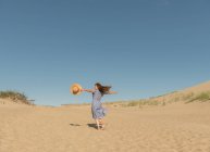 Mujer adulta en vestido casual y sombrero de paja corriendo pasar un buen rato en la duna de arena en el día de verano caliente - foto de stock