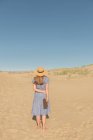 Mulher em vestido casual e chapéu de palha de pé com livro sobre duna de areia no dia quente de verão — Fotografia de Stock