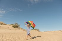 Adulto confiado mujer en vestido casual llevando arco iris bandera de color por encima de la cabeza en dunas de arena día ventoso - foto de stock