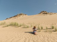 Mujer adulta reflexiva en vestido casual y sombrero de paja libro de lectura mientras está sentado en la duna de arena en el día de verano caliente - foto de stock