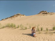 Задумчивая взрослая женщина в повседневной одежде и соломенной шляпе читает книгу, сидя на песчаной дюне в жаркий летний день — стоковое фото
