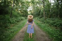 Back view of adult woman in straw hat and sundress standing along forest road between pines at sunny day — Stock Photo