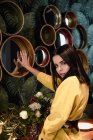 Jolie jeune femme brune à la mode dans une tenue à la mode touchant miroir à la main tout en regardant la caméra près des fleurs et des plantes à l'intérieur — Photo de stock