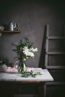 Glass vase with bunch of white flowers with leaves on marble tabletop beside dark wall — Stock Photo