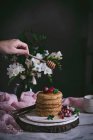 Human hand dripping honey on stack of pancakes with fresh berries on porcelain over dark background — Stock Photo