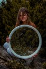 Красива жінка стоїть з великим круглим дзеркалом біля зелених кущів дивиться на камеру — стокове фото