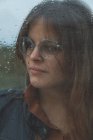 Portrait d'une belle femme avec des lunettes regardant par la fenêtre mouillée le jour de la pluie en regardant loin — Photo de stock