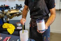 Concentrated crop man wearing dirty apron mixing various paints for serigraphy in workshop — Stock Photo