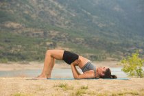 Femme adulte moyenne en pose de pont faisant du yoga en plein air sur la plage du barrage — Photo de stock