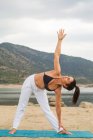 Mid adult woman in triangle pose doing yoga outdoors on dam beach — Stock Photo