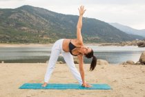 Femme adulte moyenne en pose triangle faisant du yoga en plein air sur la plage du barrage — Photo de stock