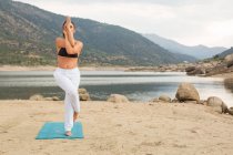 Mid adult woman in eagle pose doing yoga outdoors on dam beach — Stock Photo