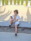 Sonriente niño casual con casco y camisa blanca sentado en el monopatín en la rampa en skatepark utilizando el teléfono móvil - foto de stock