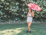 Mujer joven delgada en traje de verano con paraguas de pie piernas cruzadas cerca de árboles florecientes - foto de stock