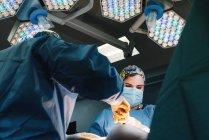Снизу серьезный молодой врач в защитной маске и кепке делает операцию с инструментами и медсестрой — стоковое фото