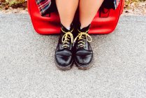 Legs of woman in black shiny boots with yellow shoelaces sitting on red suitcase waiting for transport on road — Stock Photo