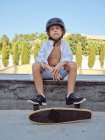 Casual criança no capacete e camisa branca sentado na rampa no skatepark olhando na câmera — Fotografia de Stock