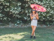 Magro jovem mulher no verão roupa com guarda-chuva pé pernas cruzadas perto florescendo árvores — Fotografia de Stock