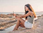 Rêveuse jeune femme tendance textos sur la plage — Photo de stock