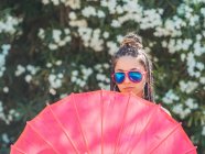 Magro jovem mulher em óculos de sol com guarda-chuva de pé perto de árvores florescentes — Fotografia de Stock