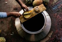 D'en haut de l'homme coupant l'ananas avec un couteau sur le dessus du réservoir en métal à la lumière du jour — Photo de stock