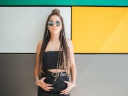 Pretty stylish teenage girl with hands in pockets and unique dreadlocks looking at camera on colorful background — Stock Photo