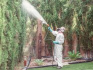 Specialist in green gloves, respiratory mask and uniform holding hose and spraying chemical on green on fence in yard — Stock Photo