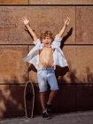 Emocionado niño gritando en camisa blanca apoyada en la pared sosteniendo los brazos de pie con monopatín en la luz del sol mirando en la cámara - foto de stock