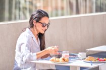 Довольно дружелюбная дама в солнечных очках сидит за столом на открытой солнечной террасе и ест розовый йогурт в ожидании партнера — стоковое фото