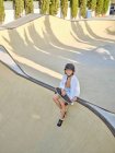 From above of smiling casual kid in helmet and white shirt sitting on ramp in skatepark looking in camera — Stock Photo