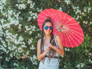 Тонка молода жінка в літньому Екіпіровка і сонцезахисні окуляри з парасолькою стоячи біля квітучих дерев — стокове фото