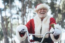 Hombre mayor en traje de Santa Claus sentado en el ciclo, timbre de campana y mirando a la cámara - foto de stock