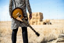 Обрежьте задний вид стильного музыканта в печатном пальто зебры с гитарой, стоящей перед забором из колючей проволоки и глядя на сухие стоги сена в поле — стоковое фото